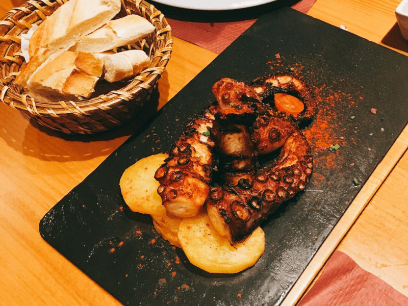 Pulpo a la parrilla con patatas en un plato negro junto a una cesta de pan, tonos cálidos