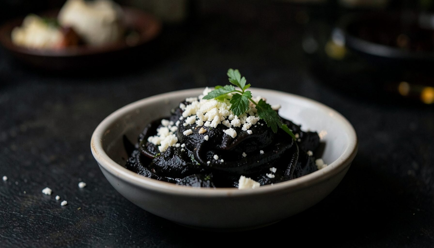 Plato de pasta negra servido en un cuenco sobre una mesa oscura, con queso rallado y hierbas frescas, iluminación tenue y ambiente gastronómico