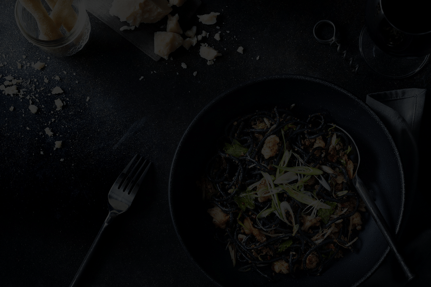 Plato de pasta negra servido en un cuenco sobre una mesa oscura, con queso rallado y hierbas frescas, iluminación tenue y ambiente gastronómico
