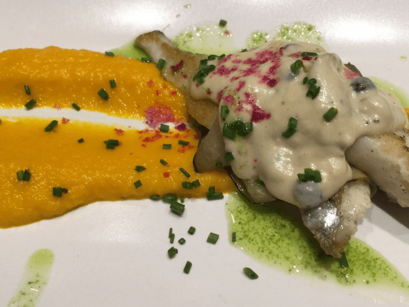 Plato de pescado emplatado con puré naranja, salsa cremosa y guarnición de hierbas verdes sobre plato blanco