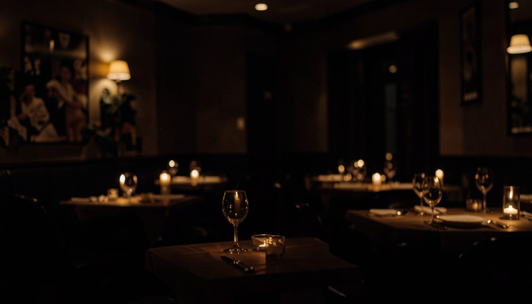 Comedor de restaurante en penumbra con mesas preparadas, copas de vino y velas, ambiente cálido y acogedor